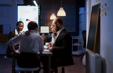 Selective focus of diverse multi-ethnic businesspeople overworking late at night analyzing management ideas solving company statistics. Workaholics teamwork working in office meeting room in evening